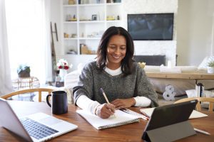 Happy mixed race woman sitting at her dining table and taking notes with a laptop and tablet open beside her.