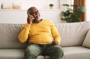 Senior man talking on phone and sitting on couch at home, and holding his glasses in one hand.